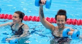 twee lachende vrouwen die aquajoggen doen in het zwembad met dumbells van foam