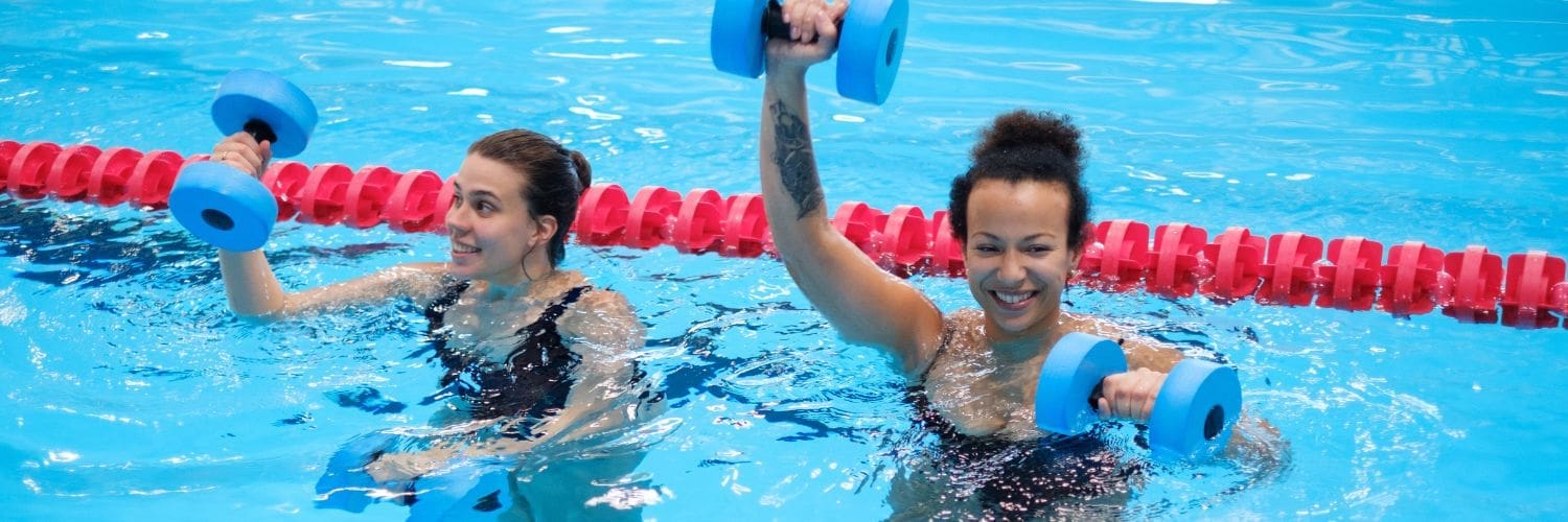 twee lachende vrouwen die aquajoggen doen in het zwembad met dumbells van foam